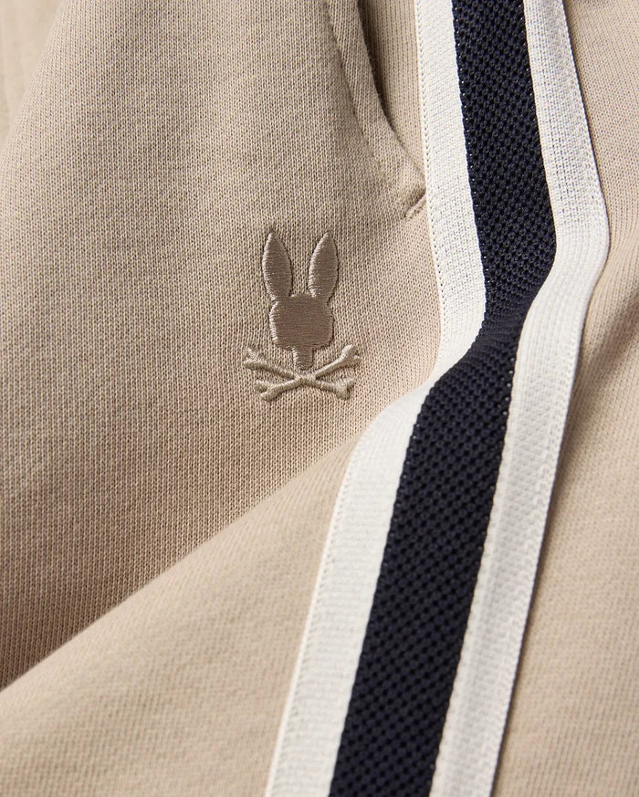 Kids Ty French Terry Sweatpant in beige french terry fabric, featuring a small embroidered Psycho Bunny skull logo and a navy and white side stripe.