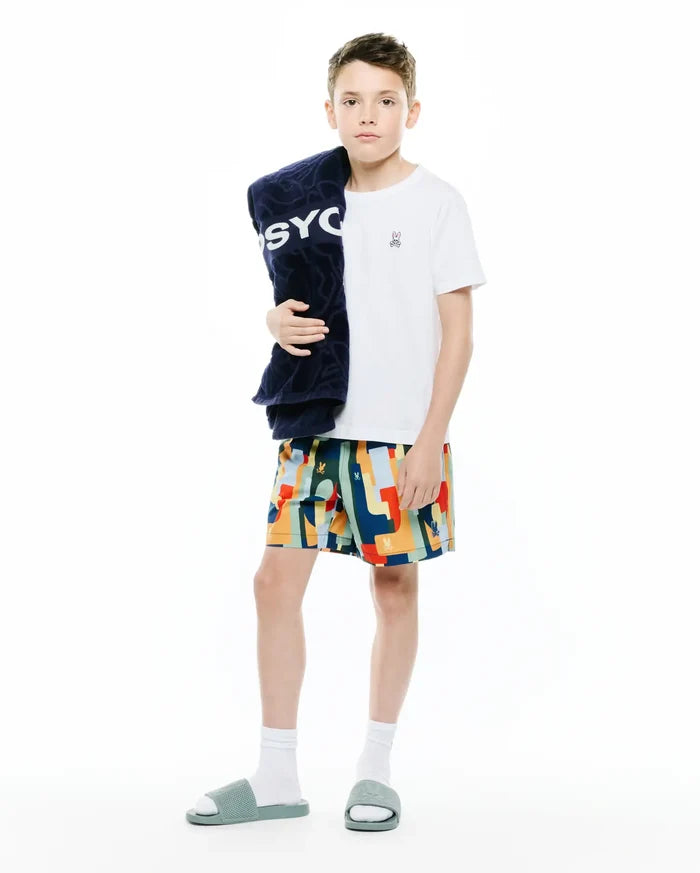 White jersey t-shirt with small embroidered Psycho Bunny logo, paired with Kids Wes All Over Print Swim Trunk featuring a vibrant geometric pattern and logo, plus a navy terry towel with large PSYCHO text.