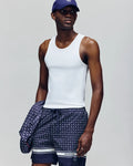 A man in a Psycho Bunny MENS 2 PACK TANK TOP - MO1400001, navy cap, and patterned swim shorts holds a matching jacket while standing against a plain grey background.