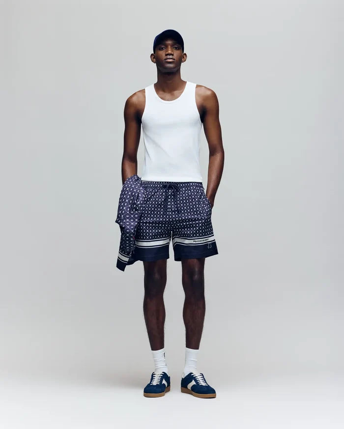 A man stands against a plain background wearing a Psycho Bunny MENS 2 PACK TANK TOP - MO1400001 in white, patterned navy shorts, a matching long-sleeve shirt tied at his waist, a navy cap, white socks, and navy sneakers.