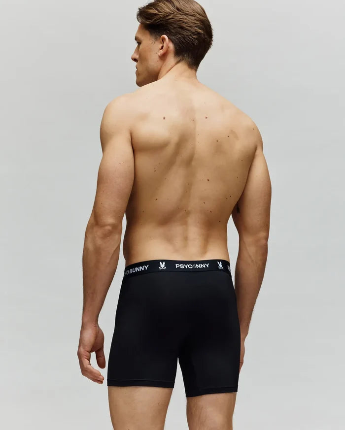 A shirtless man faces away, wearing Psycho Bunny MENS 3 PACK BOXER BRIEF - MO1400006 in black with a branded waistband. Made from a cotton-modal blend, these boxer briefs deliver all-day comfort against a neutral background.