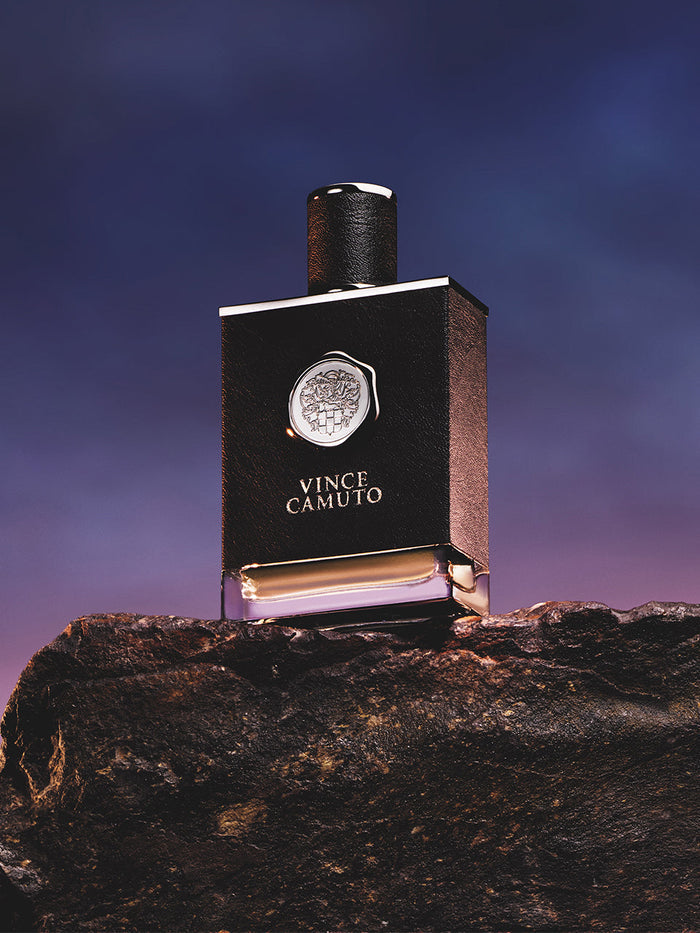 A bottle of Vince Camuto Men’s 3.4 oz Eau de Toilette Spray rests on a rugged stone, its sleek black design and silver emblem illuminated beneath a dramatic twilight sky, capturing the essence of a bold, masculine fragrance.