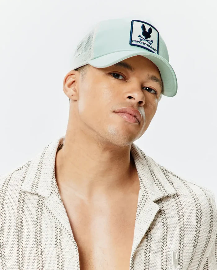 Mens Adamo Trucker Cap in mint green with white mesh and a prominent Psycho Bunny skull logo patch. Cream textured knit collared shirt with dark woven stripes and a subtle chest logo.