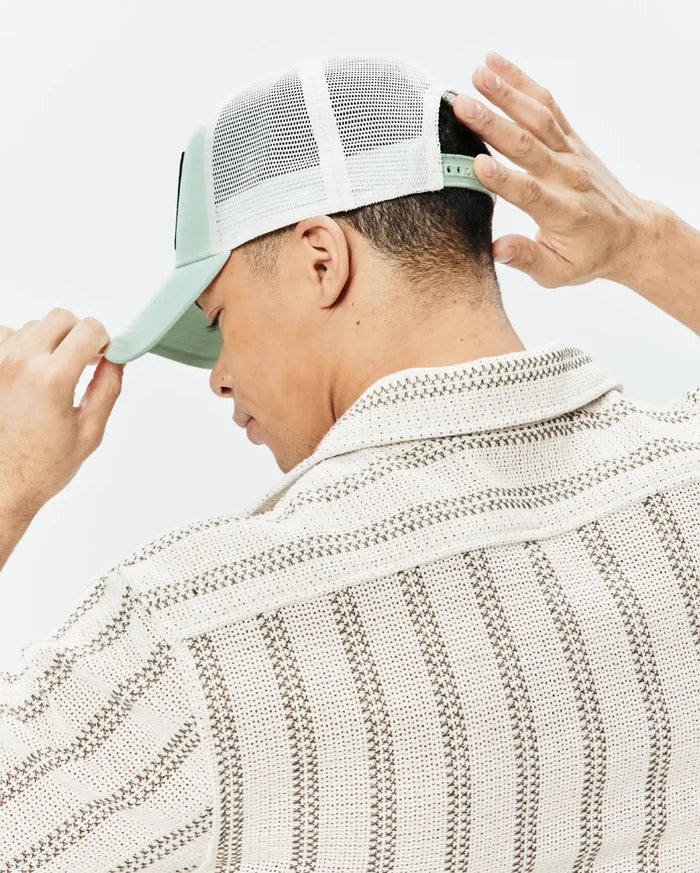 Mens Adamo Trucker Cap in mint green with white mesh back and a dark logo patch, paired with a cream textured knit short-sleeve shirt featuring brown woven stripe patterns.