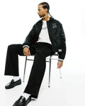 Black bomber jacket with white striped cuffs and embroidered Psycho Bunny skull logo, white jersey t-shirt with black Psycho Bunny text, and black Mens Aidan Chino Pant.