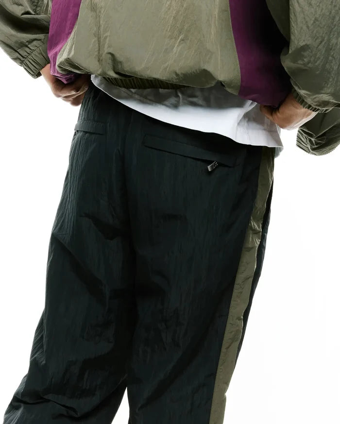 Mens Alder Nylon Colorblock Pant in deep forest green with a relaxed fit, crinkled nylon texture, and a contrasting olive green side stripe. A small zipper pull is visible on the back pocket.