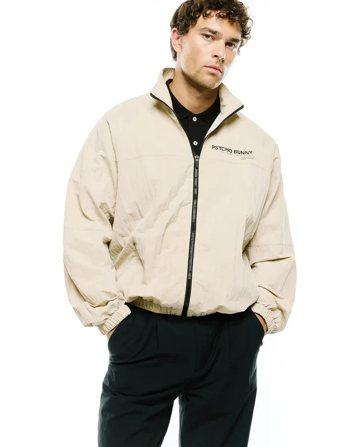 Mens Alder Nylon Jacket, a relaxed fit beige crinkled nylon full-zip jacket with a stand collar, elastic cuffs, and a black Psycho Bunny text logo with Designed For Disruption on the left chest.