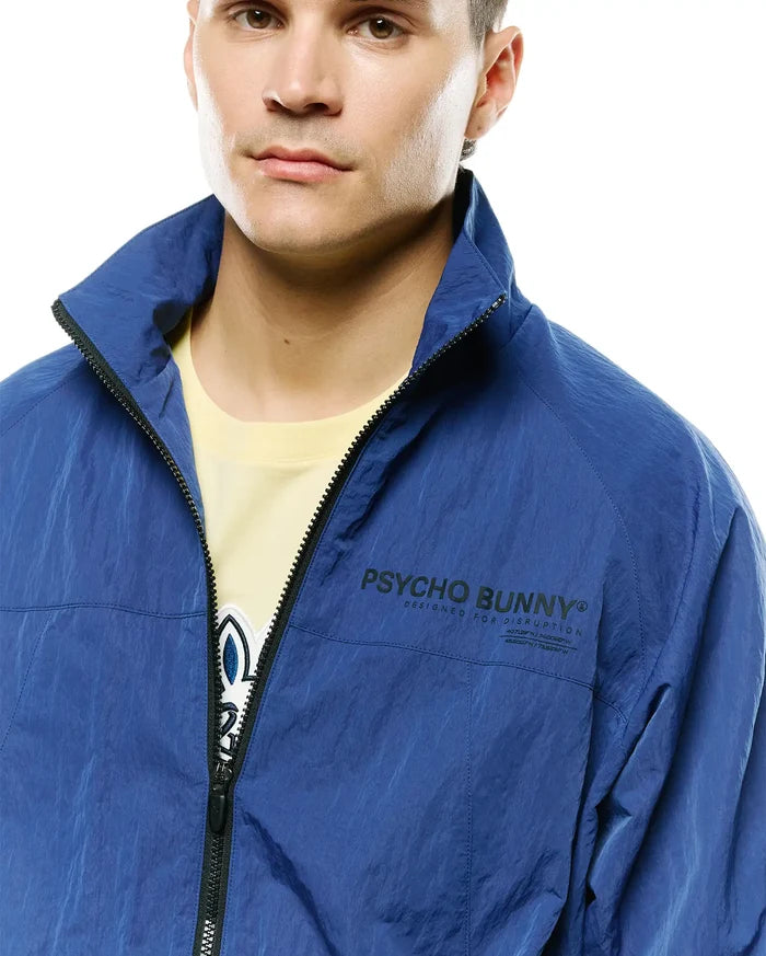 Mens Alder Nylon Jacket, royal blue crinkled nylon, relaxed fit, with a full black zipper, stand collar, and large black Psycho Bunny text logo, Designed For Disruption slogan, coordinates on left chest.