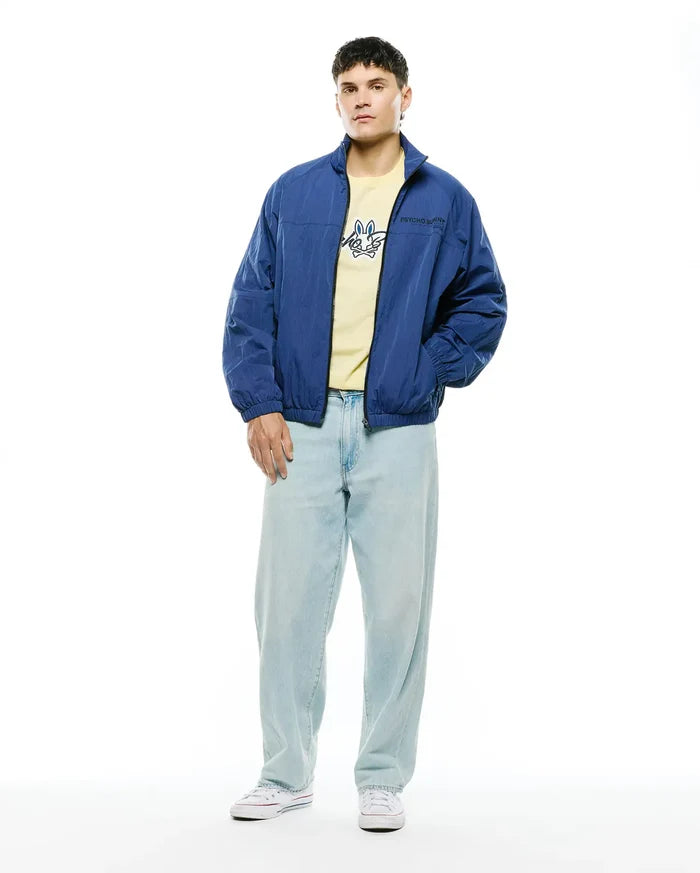 Mens Alder Nylon Jacket, a relaxed fit royal blue nylon windbreaker with a subtle Psycho Bunny chest logo, over a pale yellow jersey tee featuring a large blue bunny skull graphic, paired with light wash relaxed denim.