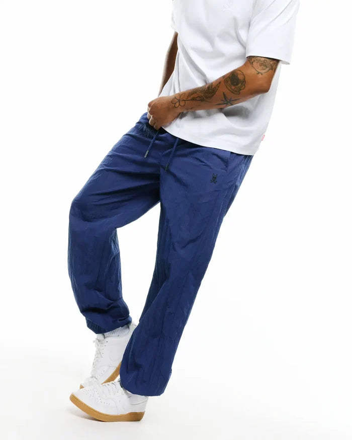 Mens Alder Nylon Pant, deep blue crinkled nylon with a relaxed fit, featuring a drawstring waist and a small embroidered black Psycho Bunny skull logo on the left thigh.