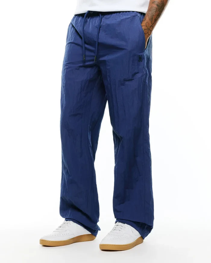 Mens Alder Nylon Pant in navy blue, featuring a relaxed fit, crinkled nylon texture, drawstring waist, and a small dark embroidered Psycho Bunny skull logo on the left thigh.