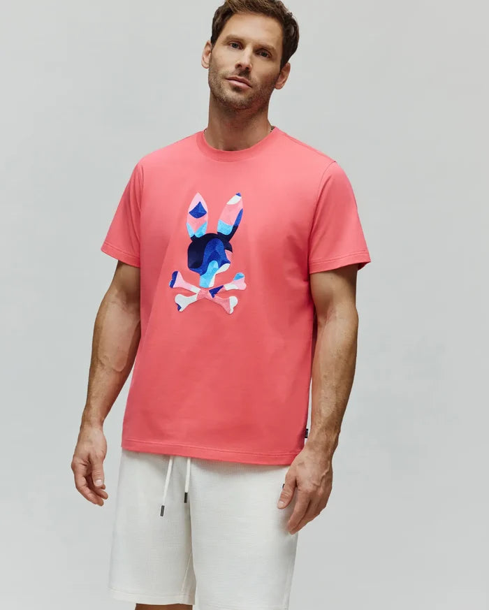 Coral pink Mens Ara Graphic Tee, classic fit jersey, with a large central Psycho Bunny skull logo featuring a vibrant blue, pink, and white camouflage pattern.