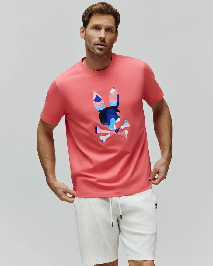 Mens Ara Graphic Tee, coral pink classic fit jersey knit t-shirt with a large, centered geometric Psycho Bunny skull logo in blue, light blue, and pink.