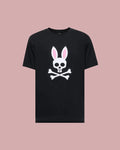 The Psycho Bunny MENS ARCHIVE BUNNY GRAPHIC TEE (MT0200031) is a black 100% Pima cotton shirt made in Peru, featuring a bold white skull with rabbit ears and pink inner accents, displayed against a light mauve background.