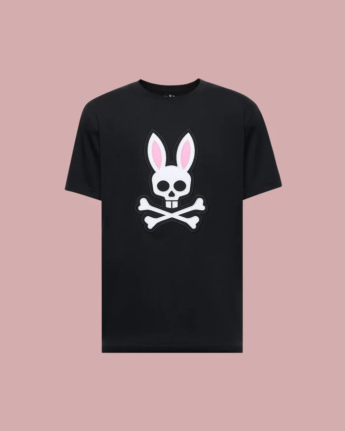 The Psycho Bunny MENS ARCHIVE BUNNY GRAPHIC TEE (MT0200031) is a black 100% Pima cotton shirt made in Peru, featuring a bold white skull with rabbit ears and pink inner accents, displayed against a light mauve background.