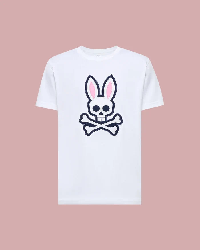 The Psycho Bunny MENS ARCHIVE BUNNY GRAPHIC TEE (MT0200031) is a white Pima cotton shirt with a bold front graphic of a skull with bunny ears and crossed bones, shown on a light pink background.