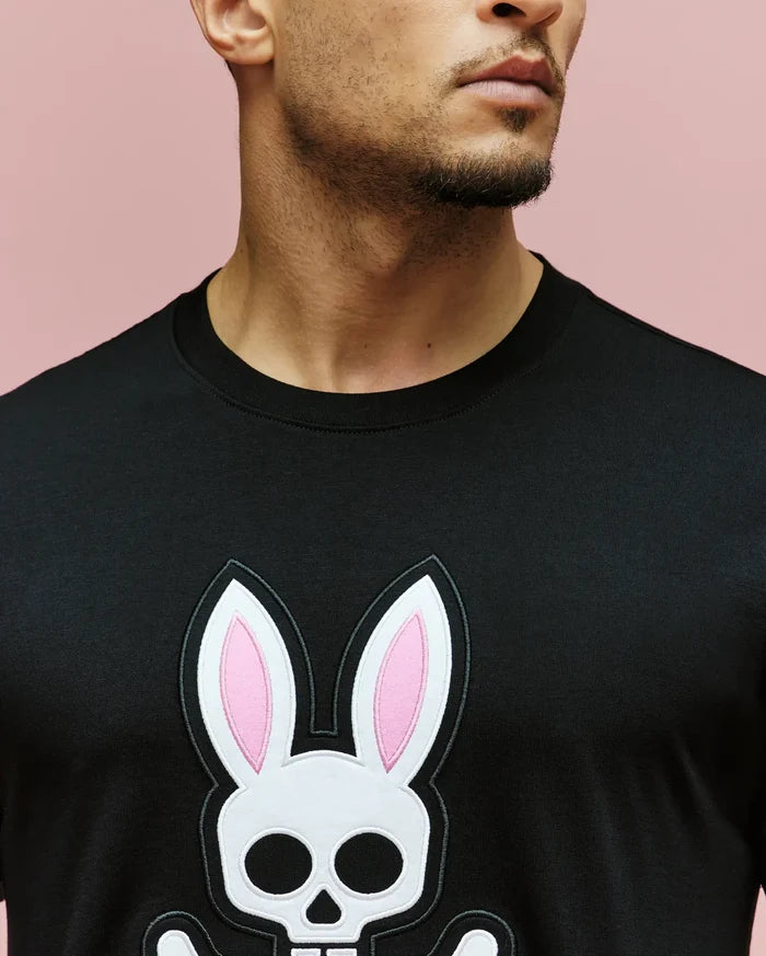 A man wears the Psycho Bunny MENS ARCHIVE BUNNY GRAPHIC TEE (MT0200031), a black 100% Pima cotton shirt with a graphic bunny skull and crossbones featuring pink-centered ears. Only his lower face and neck are visible against a light pink background.