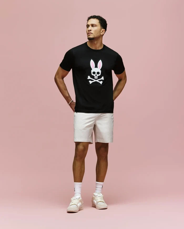 A man stands against a pink background wearing Psycho Bunny’s MENS ARCHIVE BUNNY GRAPHIC TEE (MT0200031), a black 100% Pima cotton shirt with a white skull and Embroidered Bunny with pink ears, paired with white shorts, socks, and light sneakers.