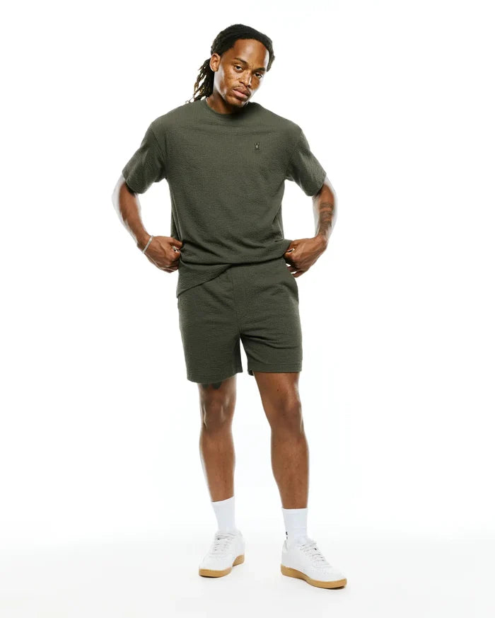 Mens Arlen Relaxed Seersucker Tee in olive green, featuring a textured seersucker fabric and a small embroidered Psycho Bunny skull logo on the left chest, paired with matching seersucker shorts.