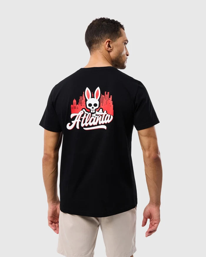 A man wearing a Psycho Bunny Black Tee crafted from Peruvian Pima cotton, featuring the MENS ATLANTA CITY TEE - B6U966X1PC graphic print with the word "Atlanta" and an emblem of a skull above crossed bones.