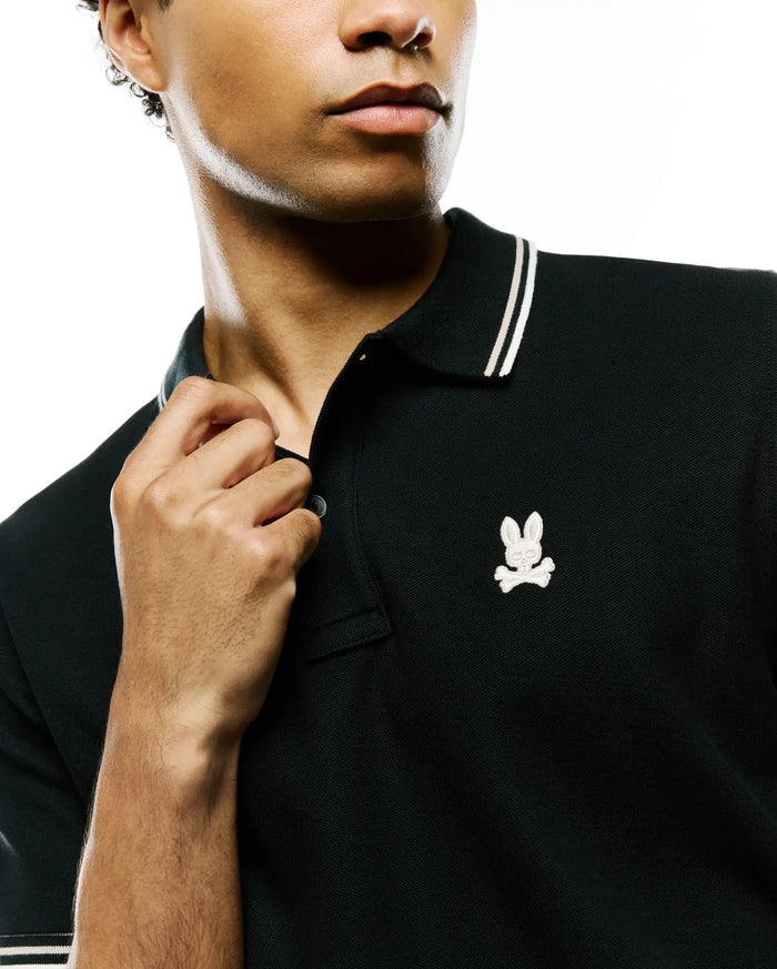 Mens Atley Pique Polo, black classic fit pique polo with white double-striped collar and cuffs, featuring a small embroidered white Psycho Bunny skull logo on the left chest.