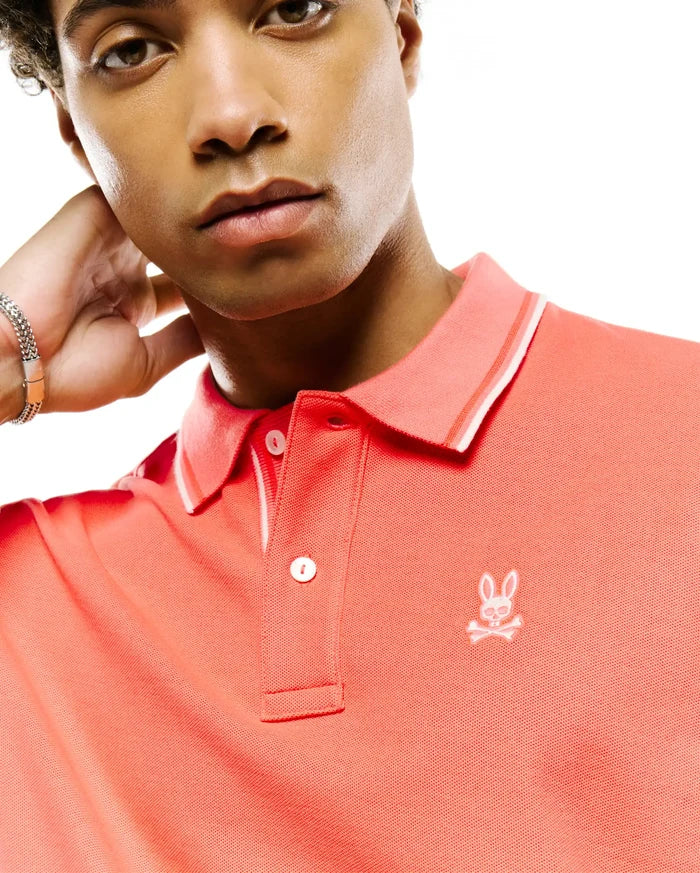 Mens Atley Pique Polo, vibrant coral, classic fit, pique knit. Features a two-button placket, ribbed collar with white tipping, and a small white embroidered Psycho Bunny skull logo on the left chest.