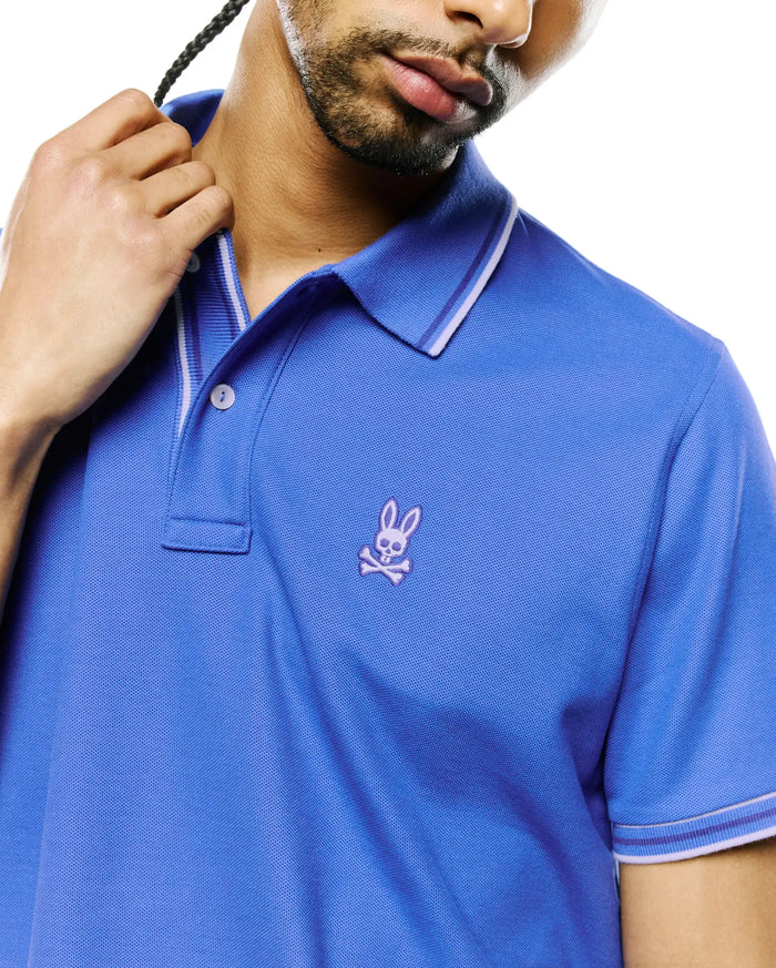 Mens Atley Pique Polo in royal blue, featuring a classic fit, pique knit texture, white and purple striped collar and cuffs, and a small embroidered purple Psycho Bunny skull logo on the left chest.