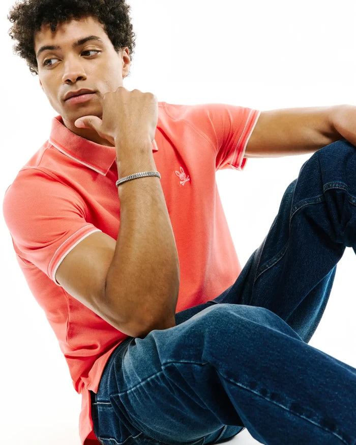 Mens Atley Pique Polo in coral, a classic fit pique knit with white tipped collar and cuffs, featuring a small embroidered Psycho Bunny skull logo on the left chest.