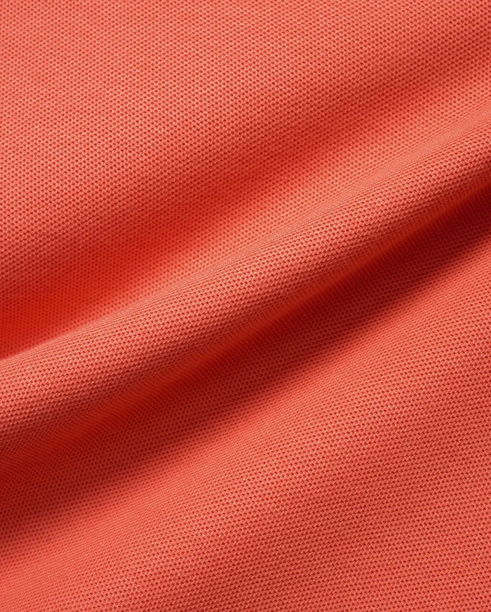 Men's Atley Pima Pique Polo in a vibrant coral orange, showcasing its premium, soft Pima cotton pique fabric with a distinct, breathable textured weave and subtle folds.