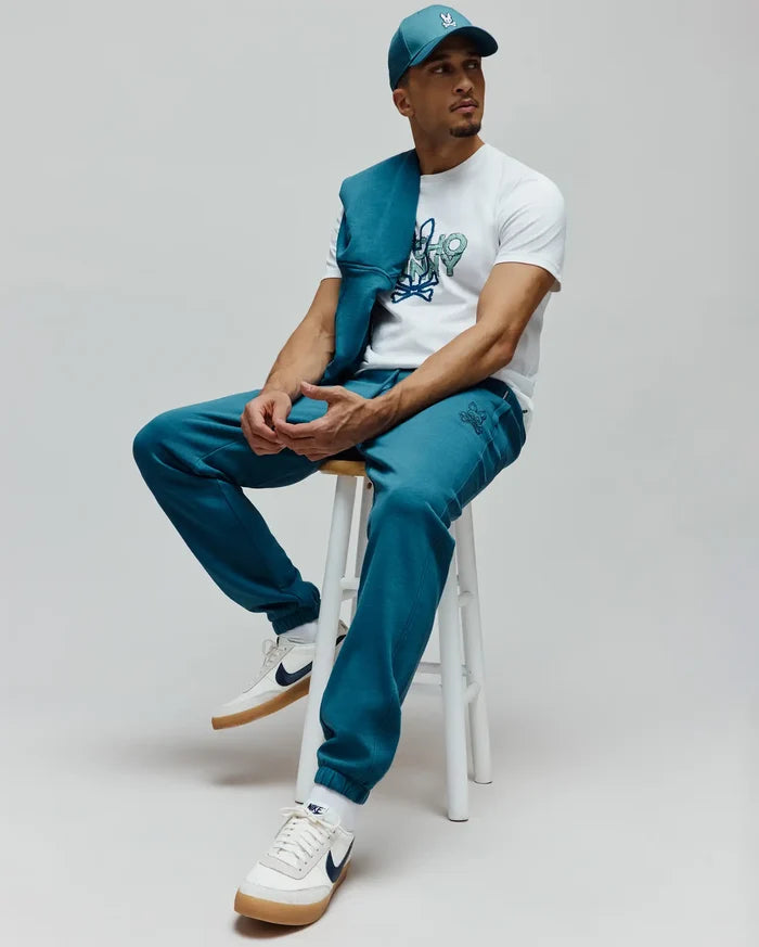 Teal cap with small embroidered Psycho Bunny skull, white classic fit jersey tee with large teal/green Psycho Bunny graphic, and relaxed fit teal French terry Mens Barry Joggers with small embroidered skull, plus a matching teal sweatshirt draped.