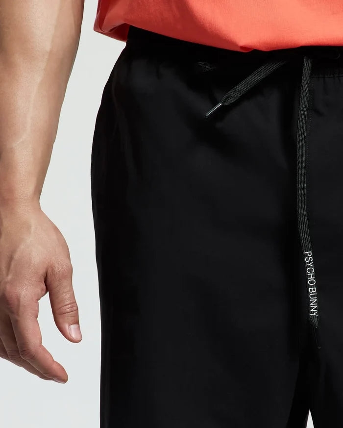 A person wears Psycho Bunny MENS BIG AND TALL SAMOS HYDROCHROMIC SWIM TRUNK with "PSYCHO BUNNY" on the drawstring and a coral shirt. The image highlights the waist area and part of the hand.