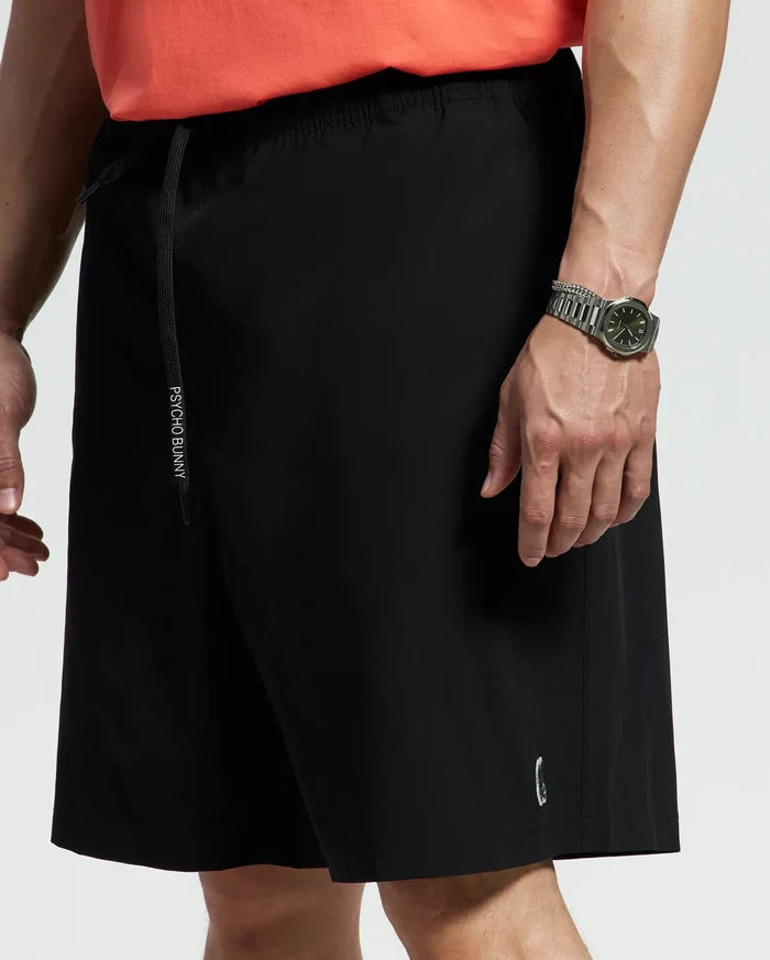 A person is seen from the waist down wearing Psycho Bunny MENS BIG AND TALL SAMOS HYDROCHROMIC SWIM TRUNK - B9W453F200 in black with a drawstring, a coral shirt, and a silver wristwatch on their left wrist. Only the lower torso and thighs are visible.