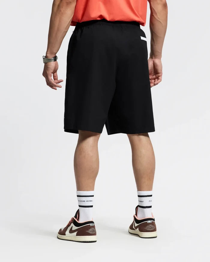 A person faces away, wearing a red shirt; Psycho Bunny MENS BIG AND TALL SAMOS HYDROCHROMIC SWIM TRUNK (black, quick-dry); white socks with black stripes; brown and white sneakers; a watch on the left wrist. The background is plain and light.