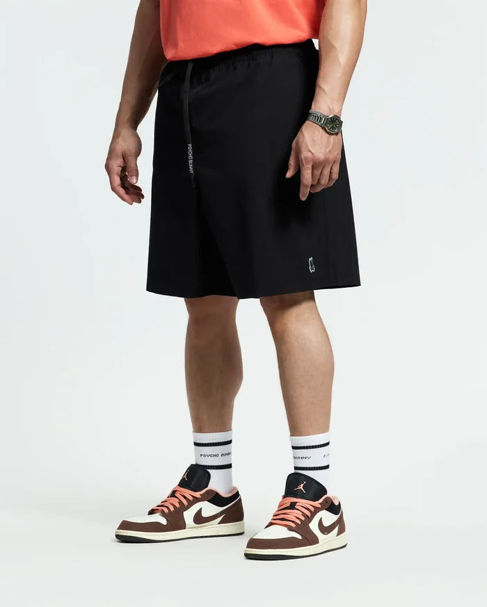 A person in coral quick-dry t-shirt, white socks, brown and white sneakers, and black Psycho Bunny MENS BIG AND TALL SAMOS HYDROCHROMIC SWIM TRUNK (B9W453F200) stands facing sideways with a watch on their relaxed left wrist.