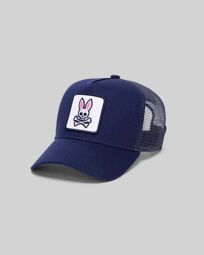 The Psycho Bunny MENS BOLD DAD TRUCKER CAP (MO2600001) is a navy blue cap with a curved brim, mesh sides and back, and features a front patch showing a pink-eared bunny above crossed bones on a white background.
