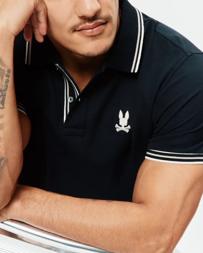 Mens Brad Pique Polo, a classic fit navy pique polo shirt with white double-striped collar and sleeve cuffs, featuring a small embroidered white Psycho Bunny skull logo on the left chest.