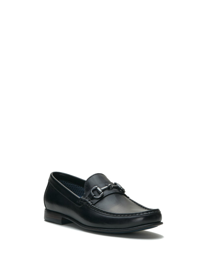 The Vince Camuto Men’s Caelan Loafer is a single black leather shoe featuring horse-bit hardware on the upper, a moccasin toe, and a low heel, displayed against a white background.