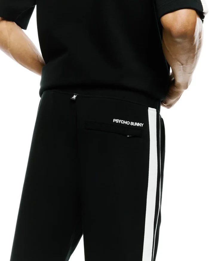 Mens Ceano Track Pant, black smooth knit with a white side stripe, featuring a white Psycho Bunny skull tag on the back waist and embroidered PSYCHO BUNNY text above the back pocket.