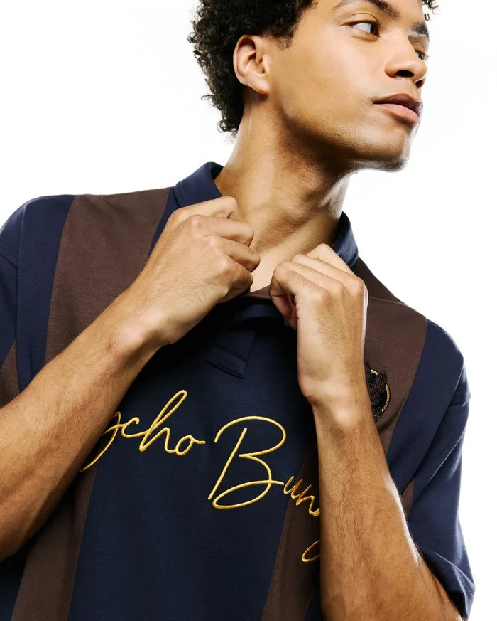 Mens Chad Relaxed Fit Striped Polo, navy blue and brown vertical striped jersey knit with a large gold embroidered Psycho Bunny script logo on the chest and a small dark bunny skull patch.