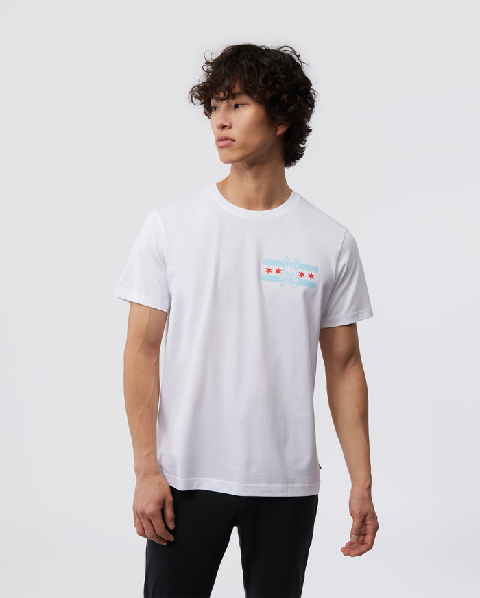 A person with curly hair is wearing the Psycho Bunny MENS CHICAGO CITY TEE - B6U551W1PC, crafted from Pima cotton jersey. The shirt features a subtle design with red stars and a light blue symbol on the chest. They are standing against a plain gray background, facing slightly to the right.