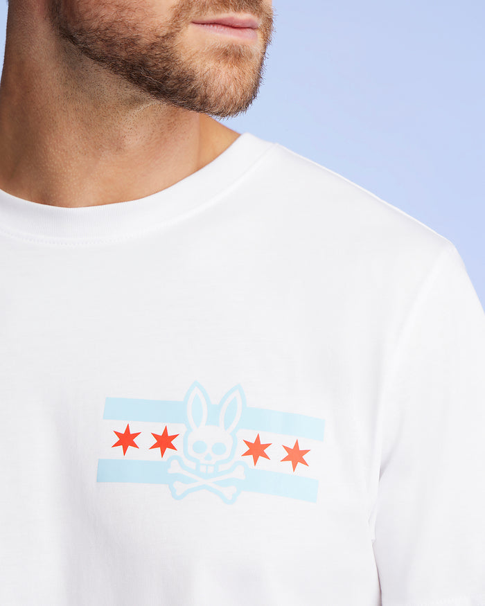 The man wears the Psycho Bunny Men's Chicago City Tee - B6U551W1PC, made from soft Pima cotton jersey. This tee features a striking blue and red design with skull elements, rabbit ears, and stars on a light blue background, perfectly highlighting the quirky Bunny logo graphic.