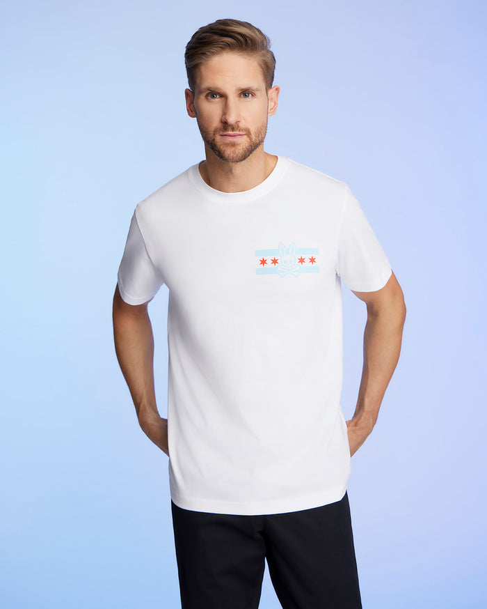 A man with short hair and a beard is wearing the Psycho Bunny MENS CHICAGO CITY TEE - B6U551W1PC, crafted from soft Pima cotton jersey. This white tee features a small design of blue stripes and red stars on the chest. He stands against a light blue background.