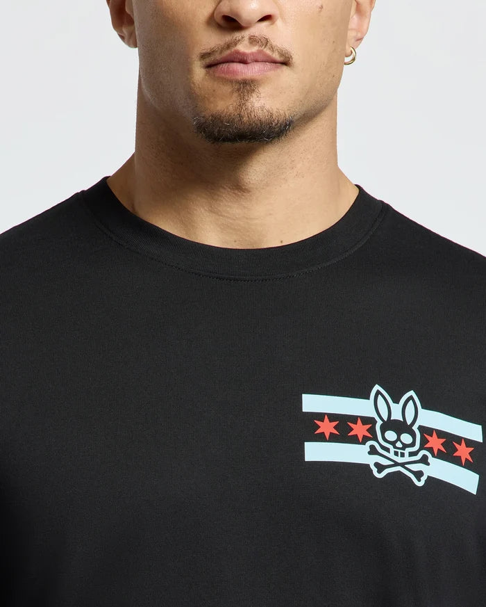 A person wearing a stylish Men's Chicago Tee by Psycho Bunny in black, featuring a graphic design of a bunny skull and crossbones surrounded by blue stripes and red stars. The image highlights the upper portion of this shirt.