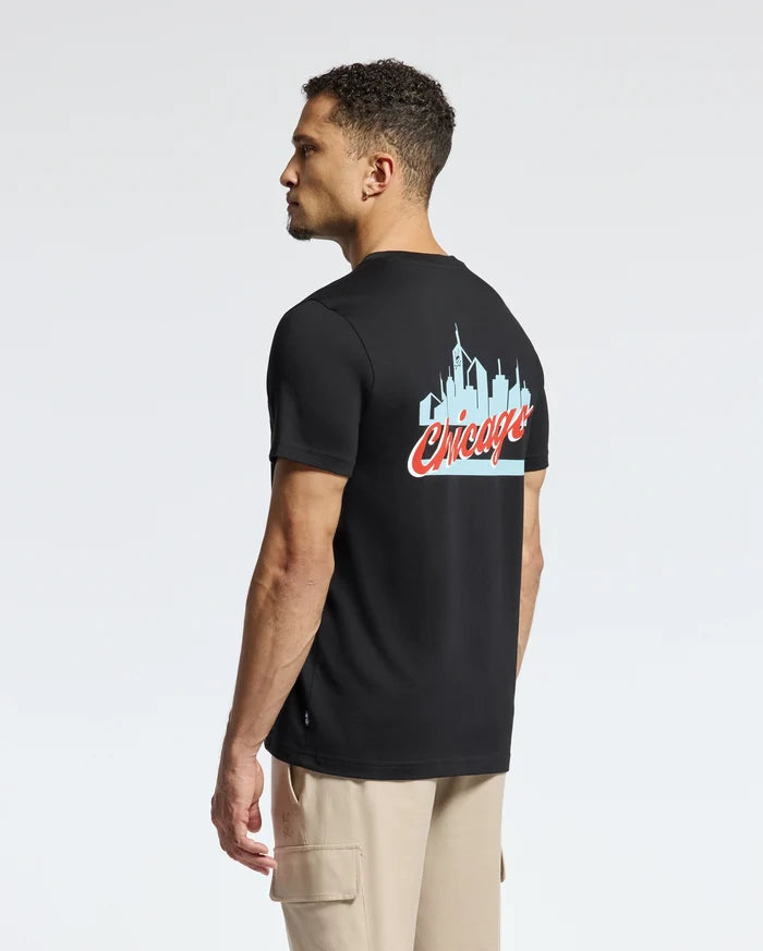 A man wearing a Psycho Bunny MENS CHICAGO TEE - B6U551W1PC crafted from soft Pima cotton is seen standing sideways against a plain background. The black tee prominently showcases a light blue skyline graphic with "Chicago" written in red cursive on the back, paired with beige cargo pants to complete his look.