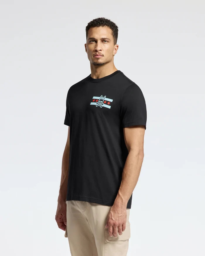 A man is standing against a plain background, wearing the Psycho Bunny MENS CHICAGO TEE - B6U551W1PC in black. This Pima cotton shirt features a small logo on the chest with a cartoonish bird and star design. He has short hair and looks directly at the camera, pairing it with light-colored pants.