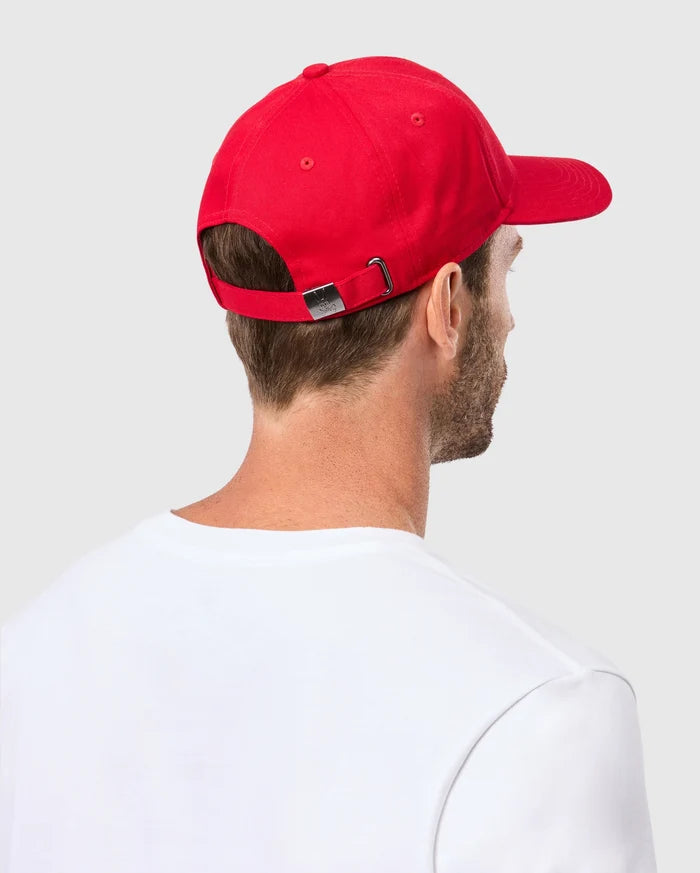 From behind, a person is seen wearing the Psycho Bunny MENS CLASSIC BASEBALL CAP - B6A816A1HT in red cotton twill with an adjustable fit, paired with a white T-shirt against a light gray background.