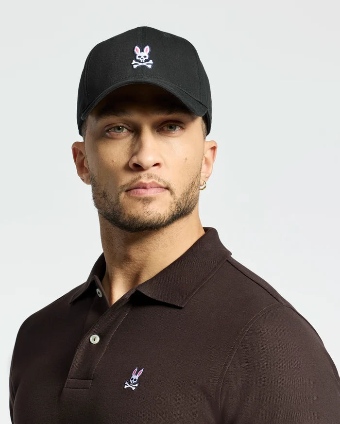 A person dons the MENS CLASSIC BASEBALL CAP by Psycho Bunny, featuring an embroidered Bunny logo. They pair it with a dark polo, possess a short beard and an earring, and pose against a light gray background, highlighting the adjustable fit of both items for comfort.