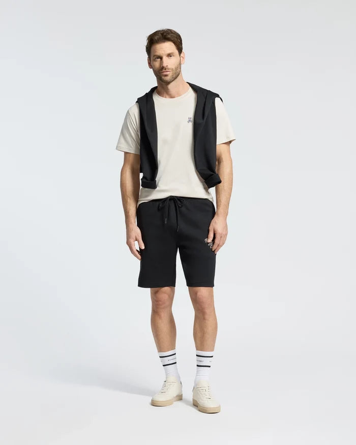 A man stands wearing a Psycho Bunny MENS CLASSIC CREW NECK TEE in white, paired with black shorts and a black jacket tied around his shoulders. He completes the look with striped socks and white sneakers against a plain white background.