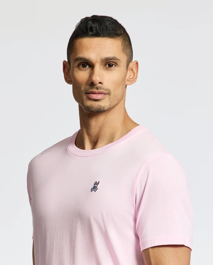 A man with short black hair poses against a plain background, wearing the versatile Psycho Bunny MENS CLASSIC CREW NECK TEE in light pink, made from soft pima cotton and featuring a small embroidered emblem.