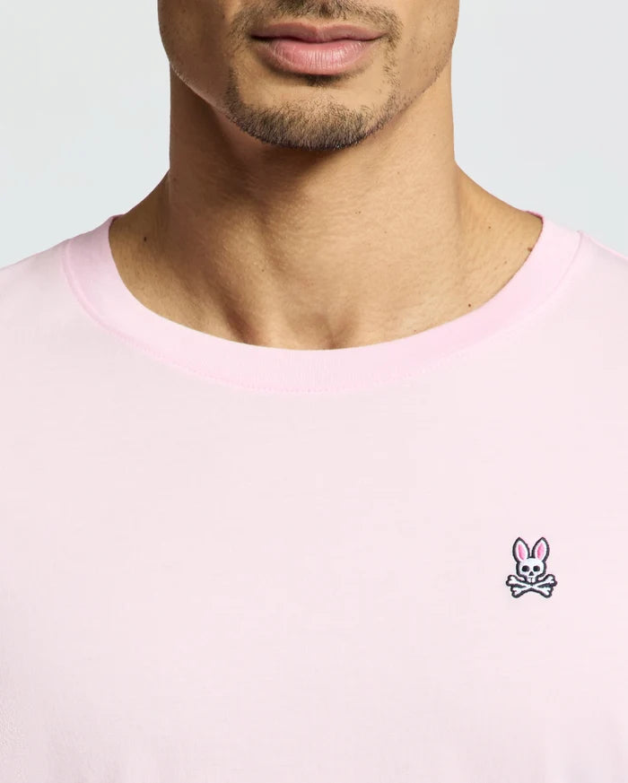 A person is shown from shoulders to just above the mouth, wearing a Psycho Bunny MENS CLASSIC CREW NECK TEE - B6U014CRPC in versatile light pink, made from soft Pima cotton with a classic crewneck and a small embroidered chest logo of a bunny face over crossed bones.